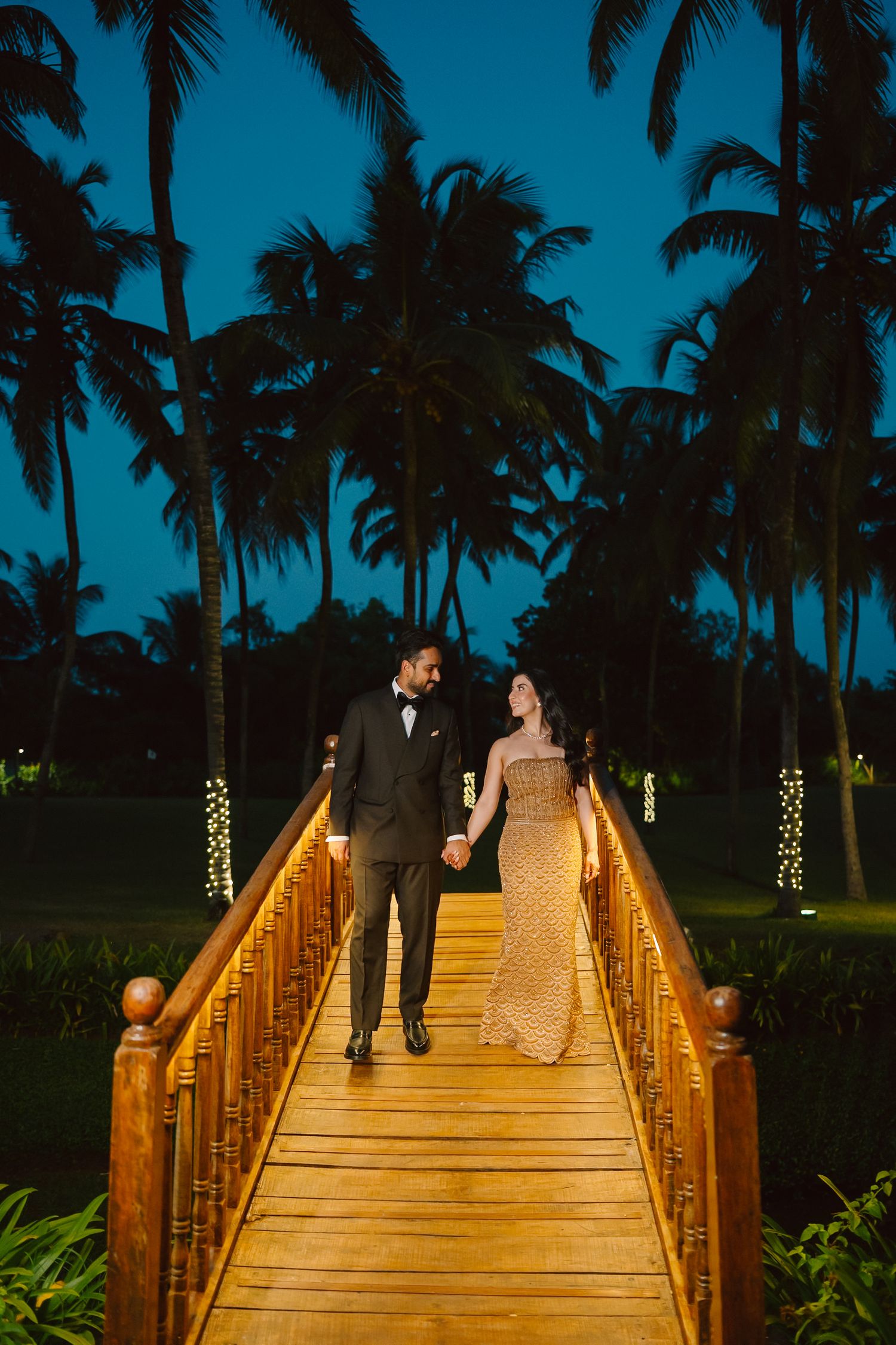 A romantic evening scene on an illuminated wooden bridge surrounded by palm trees.