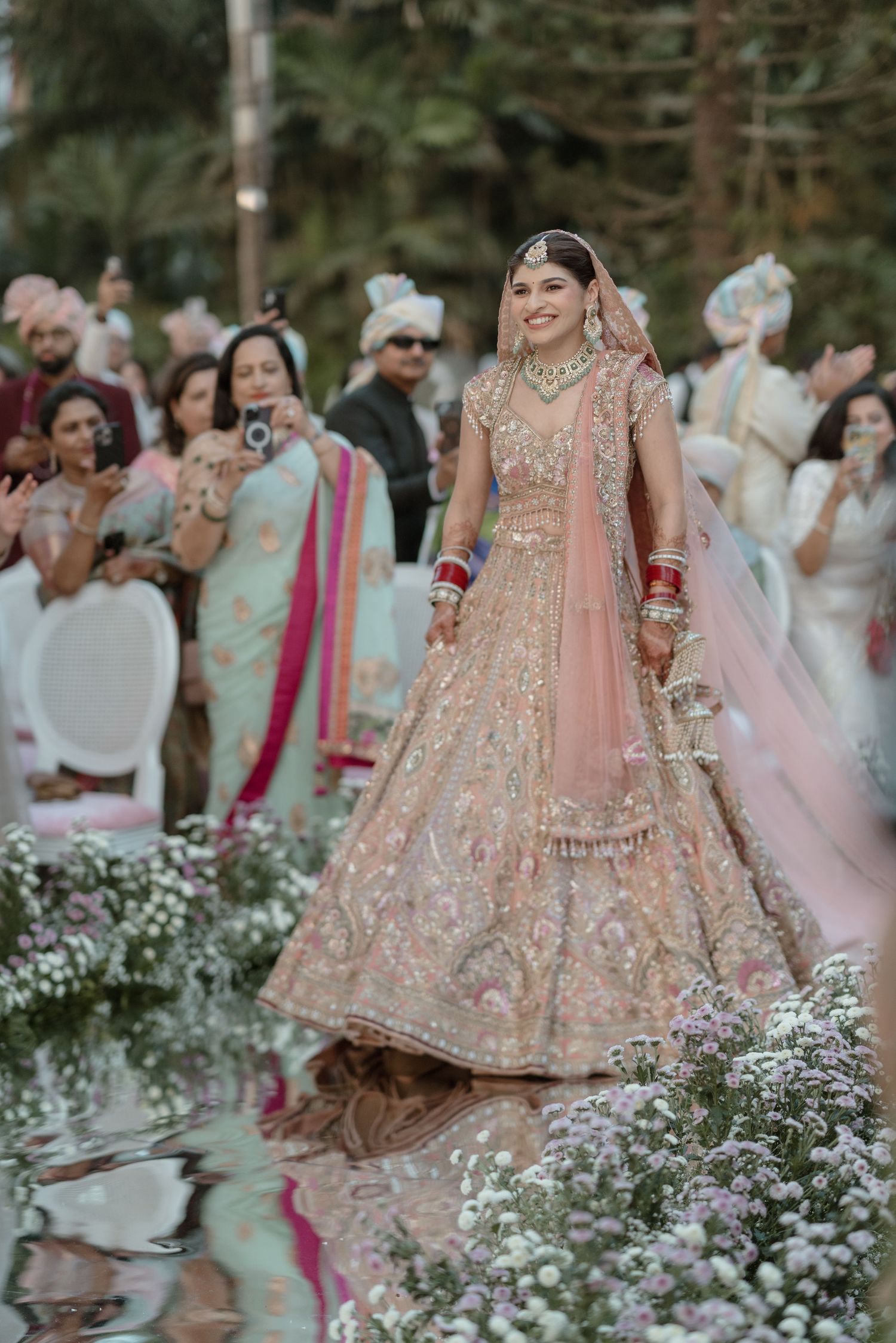 A series of outdoor wedding photos showing a bride in a pink embellished lehenga surrounded by white flowers.