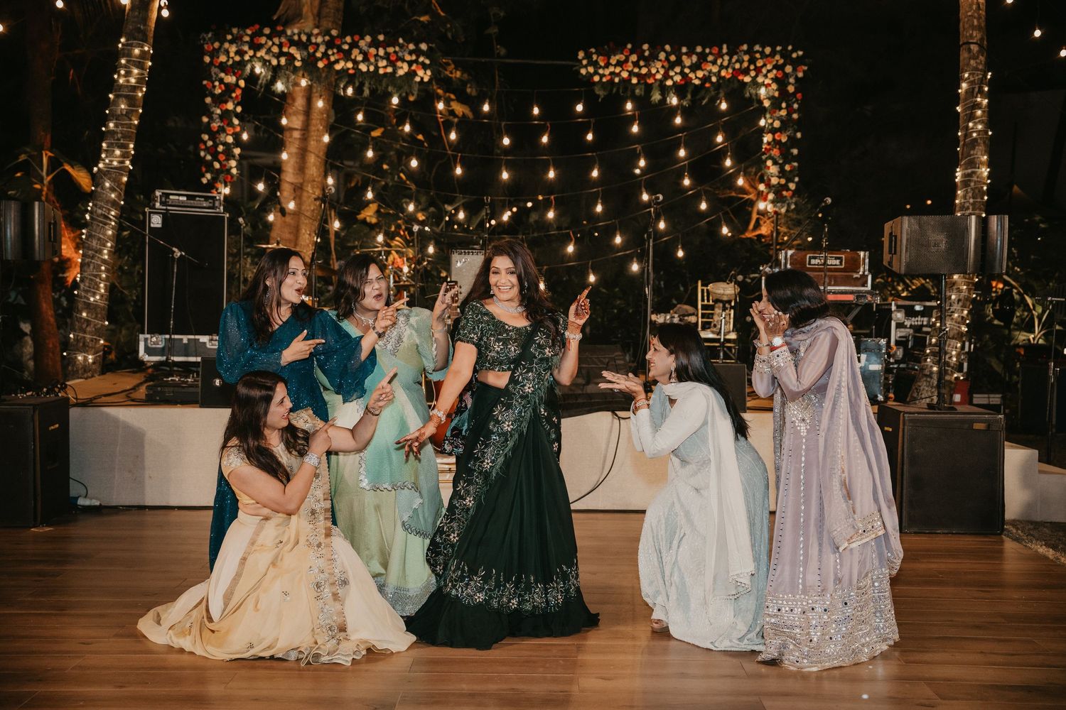Women in elegant Indian attire dancing at a festive wedding celebration with warm string lights backdrop.