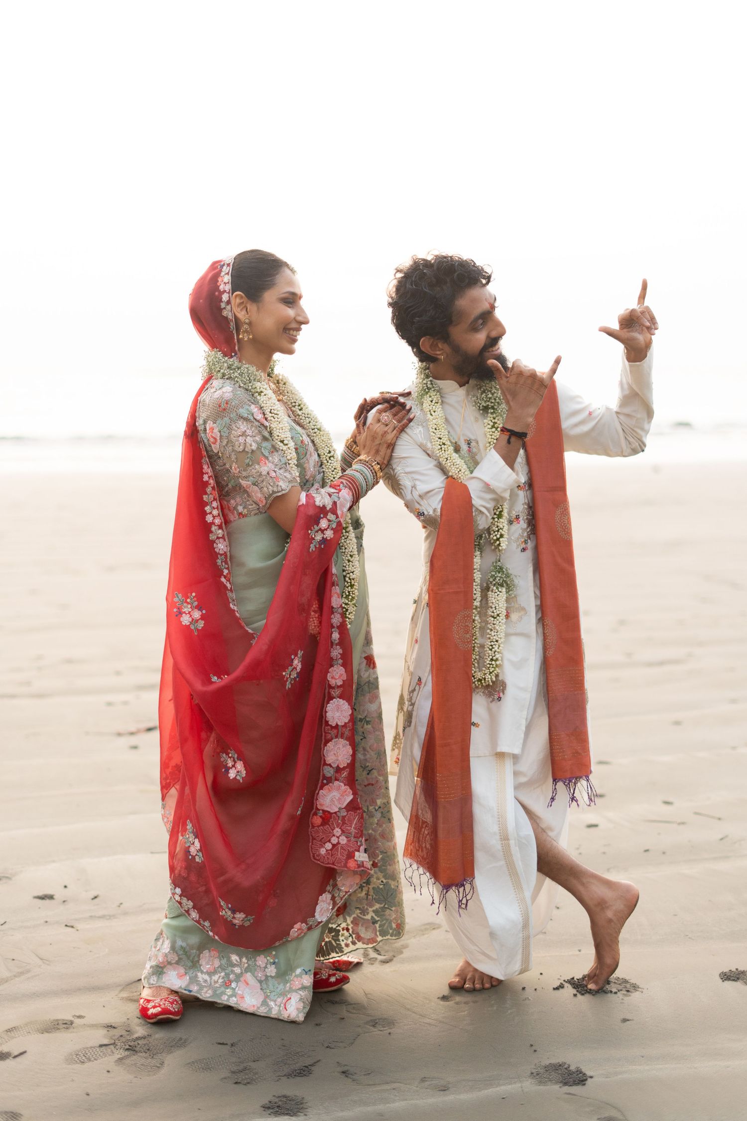 Indian couple in traditional wedding attire walking on a beach, woman in red saree, man in white kurta with floral garlands.