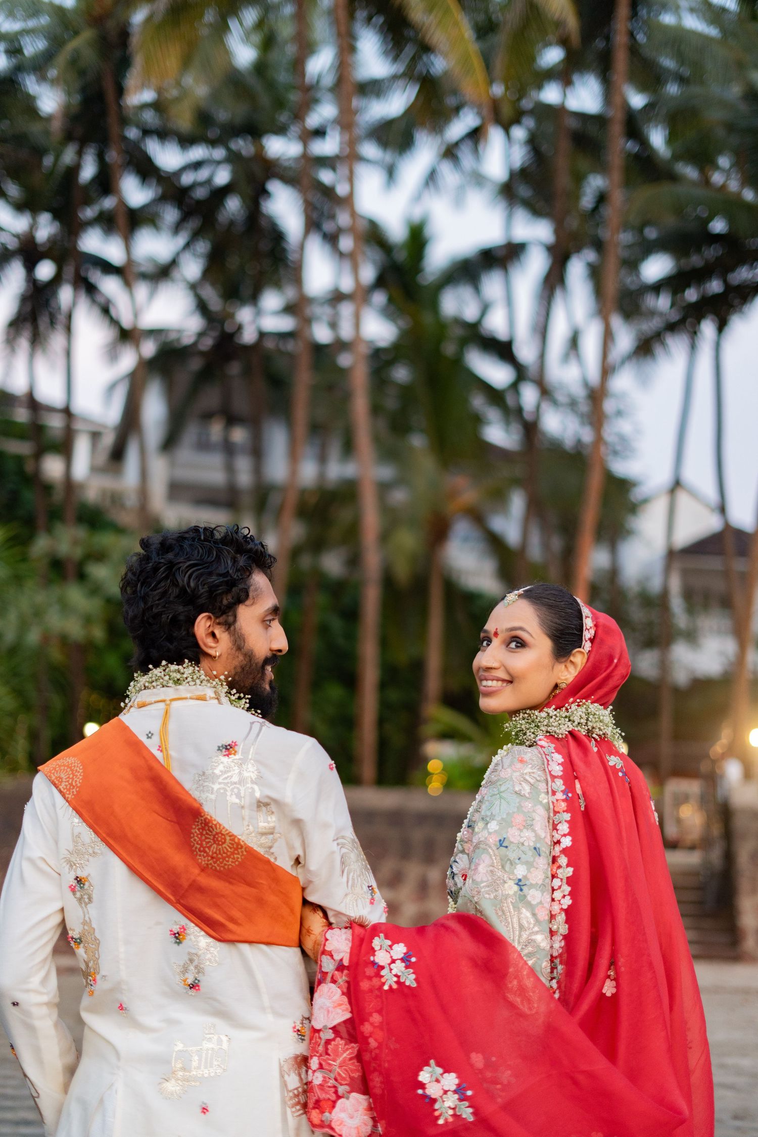 Indian couple in traditional wedding attire with floral garlands, posing outdoors with palm trees in background.