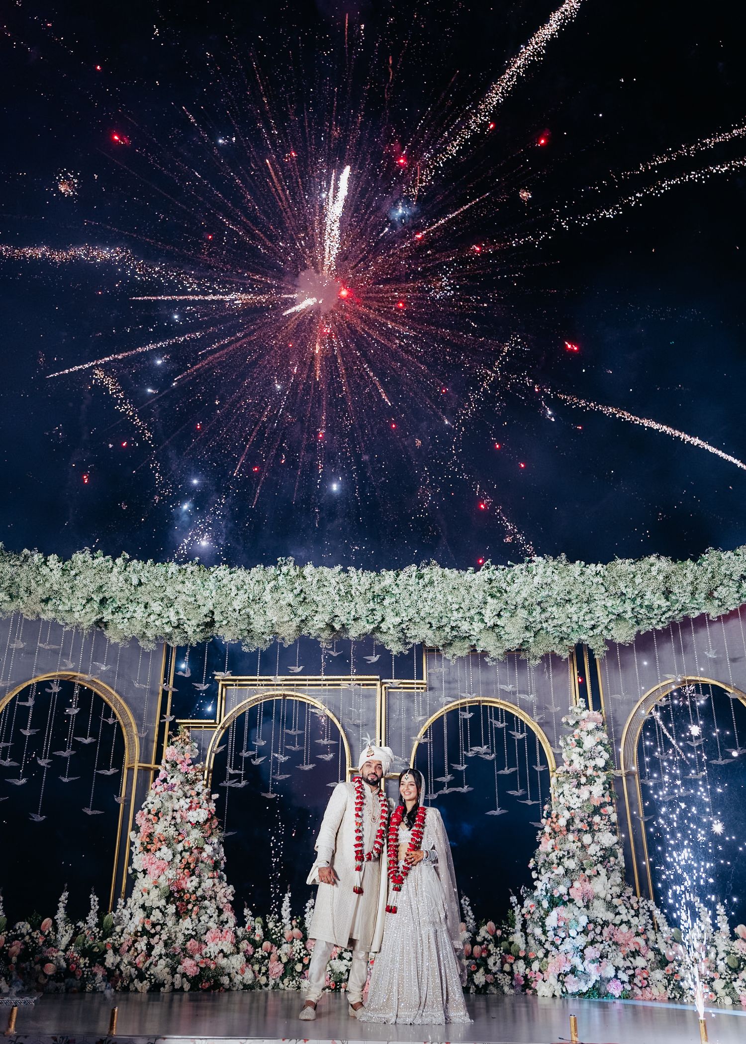A wedding celebration with fireworks lighting up the night sky above floral decorations.