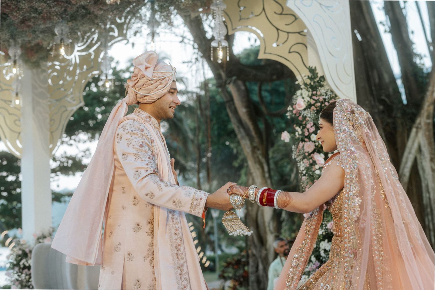 Bride in a peach-colored lace gown poses elegantly in a garden setting with natural greenery.