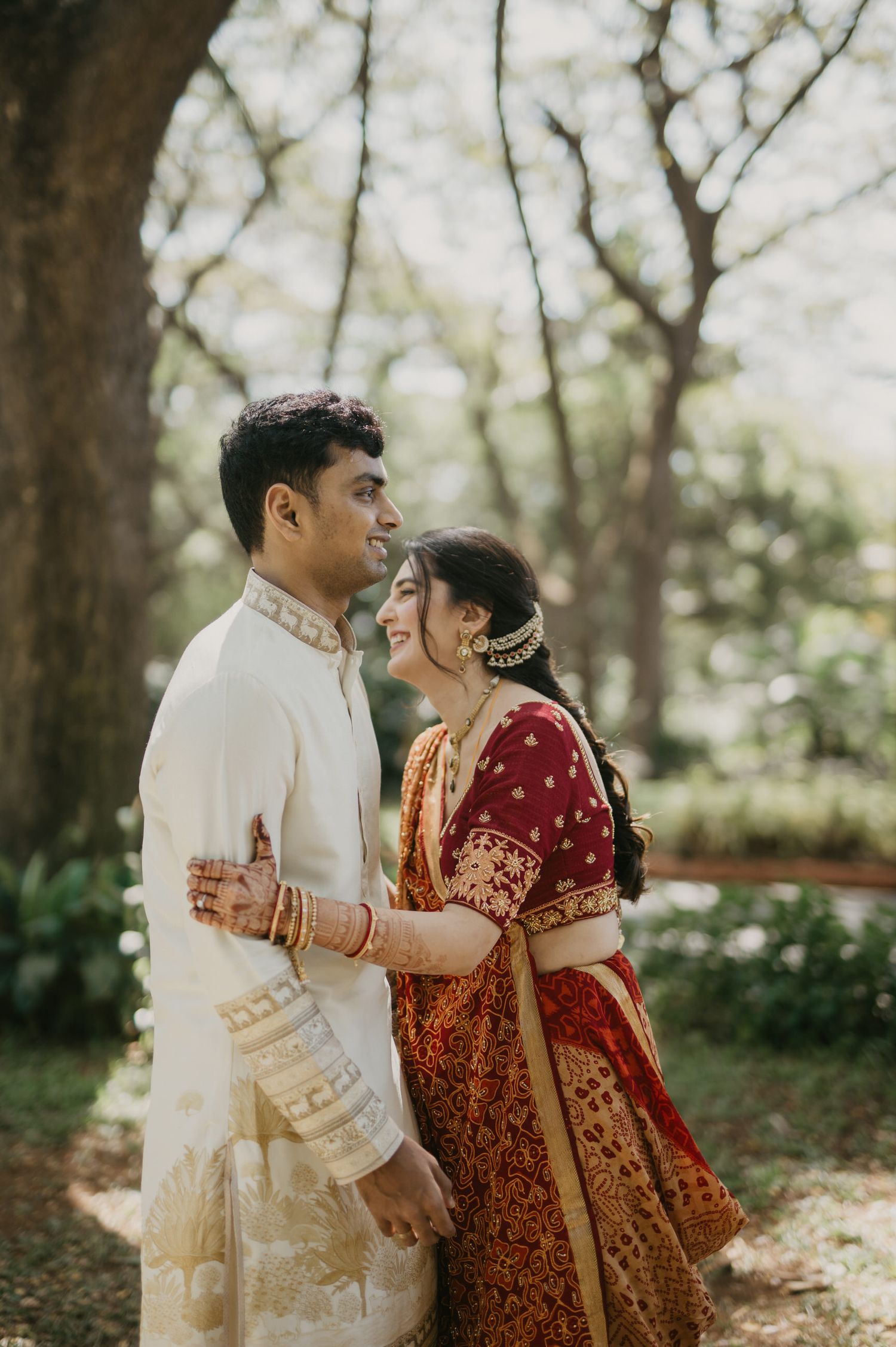 A couple shares a tender moment in traditional Indian wedding attire beneath trees in a garden setting.