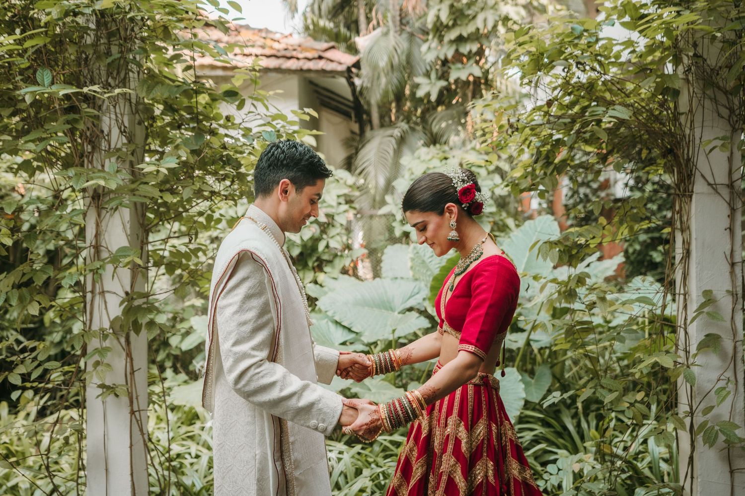 Two people in formal attire share a tender moment in a lush garden setting framed by greenery and white walls.