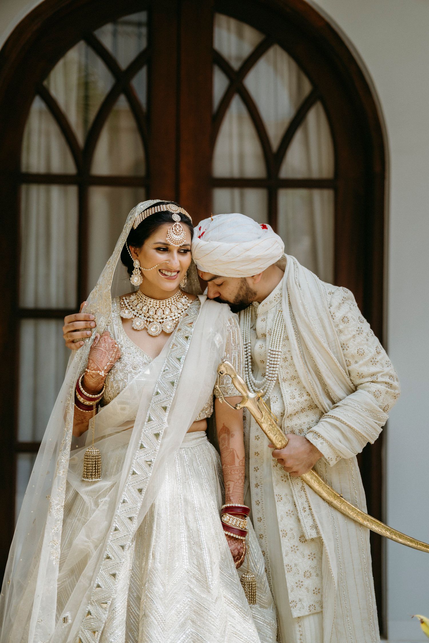 Elegant couple in matching white traditional Indian wedding outfits pose against arched doorways.