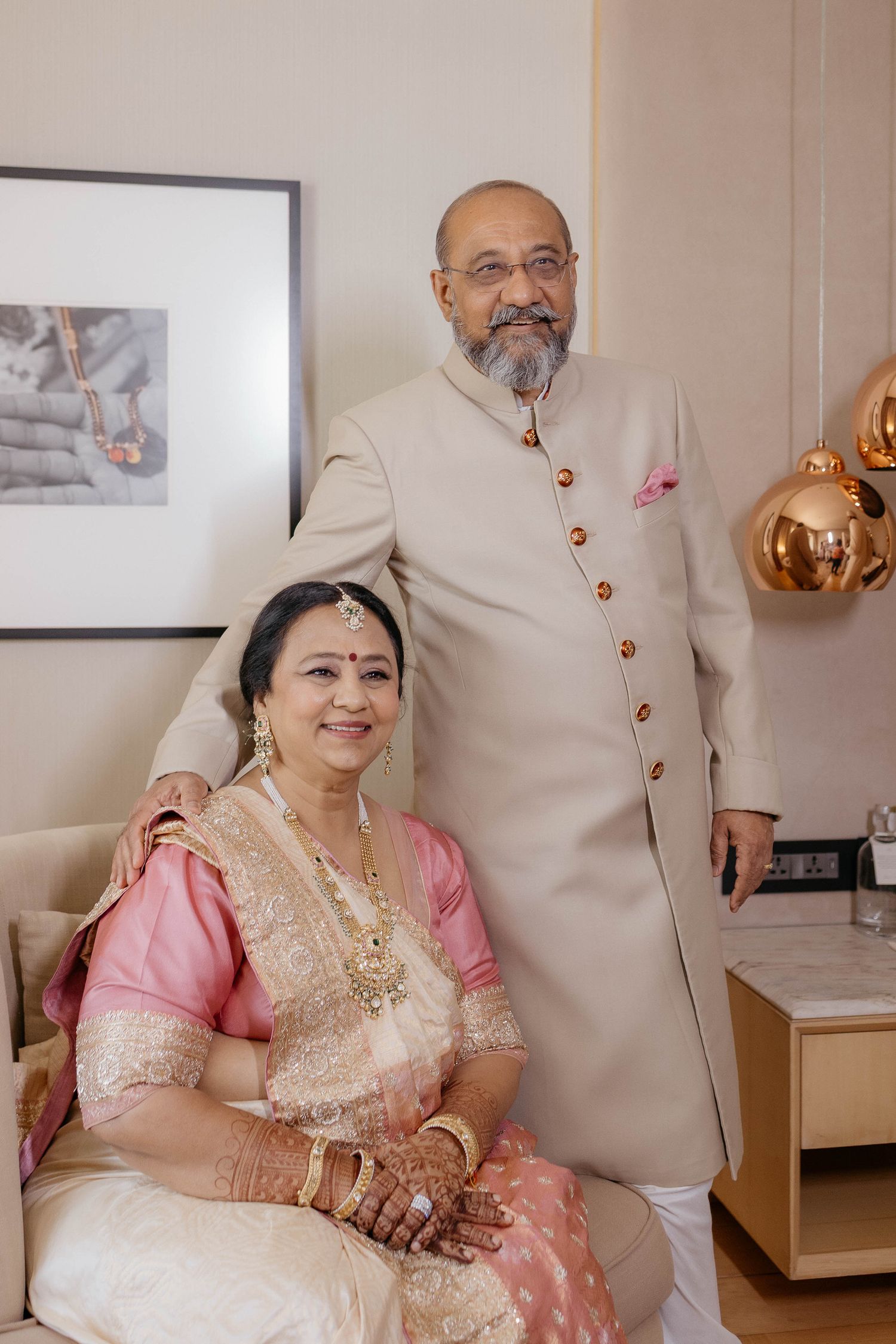 Couple in traditional Indian attire poses together in an elegant interior setting with pink gold decor details.