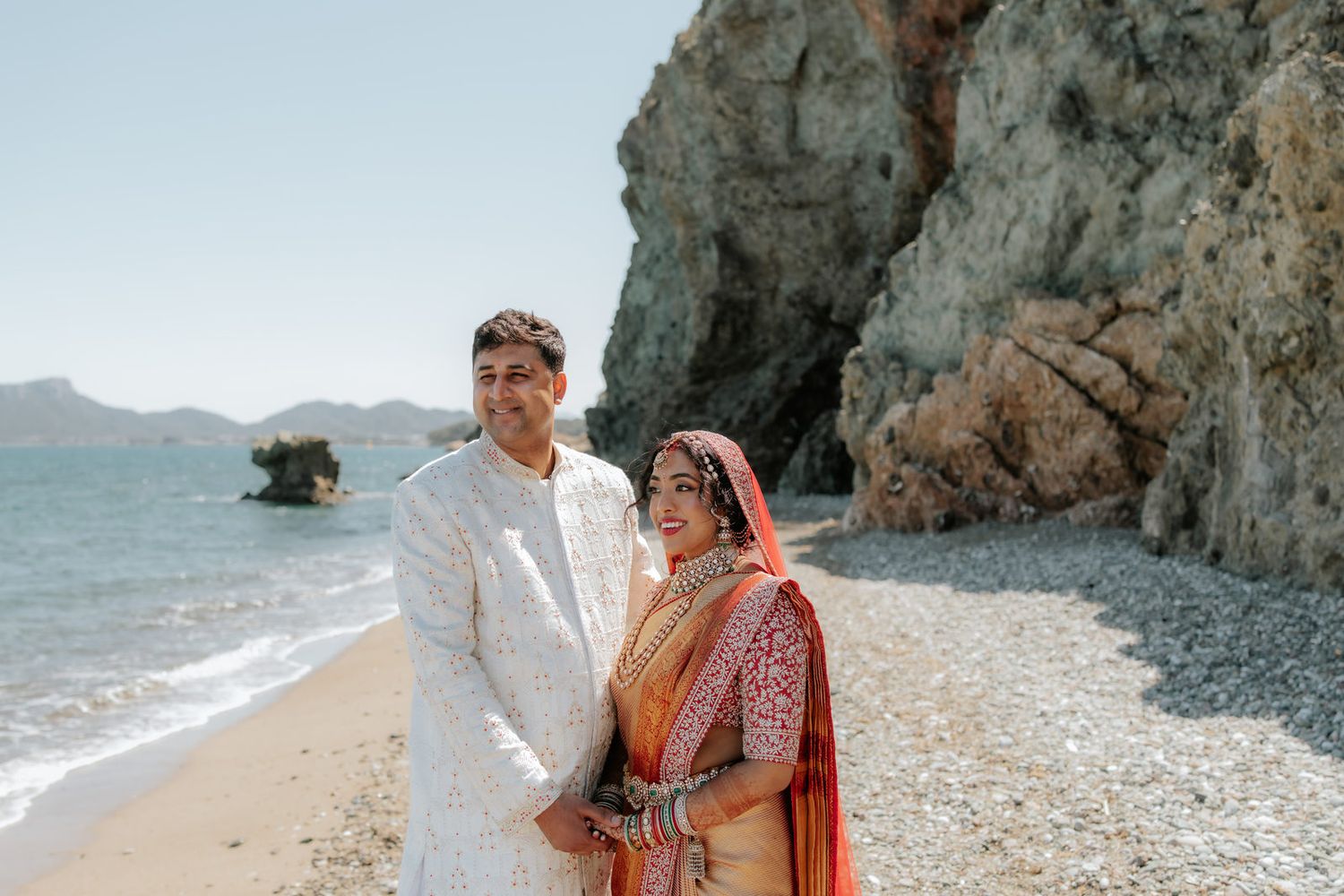 Indian couple in traditional wedding attire posing on a rocky beach, bride in red saree and groom in white sherwani.