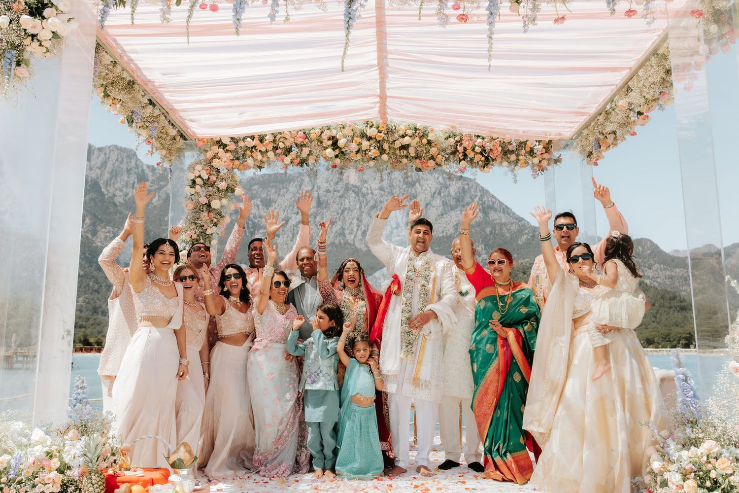 Joyful wedding party celebrates under floral mandap with mountain lake backdrop, bride and groom surrounded by family.