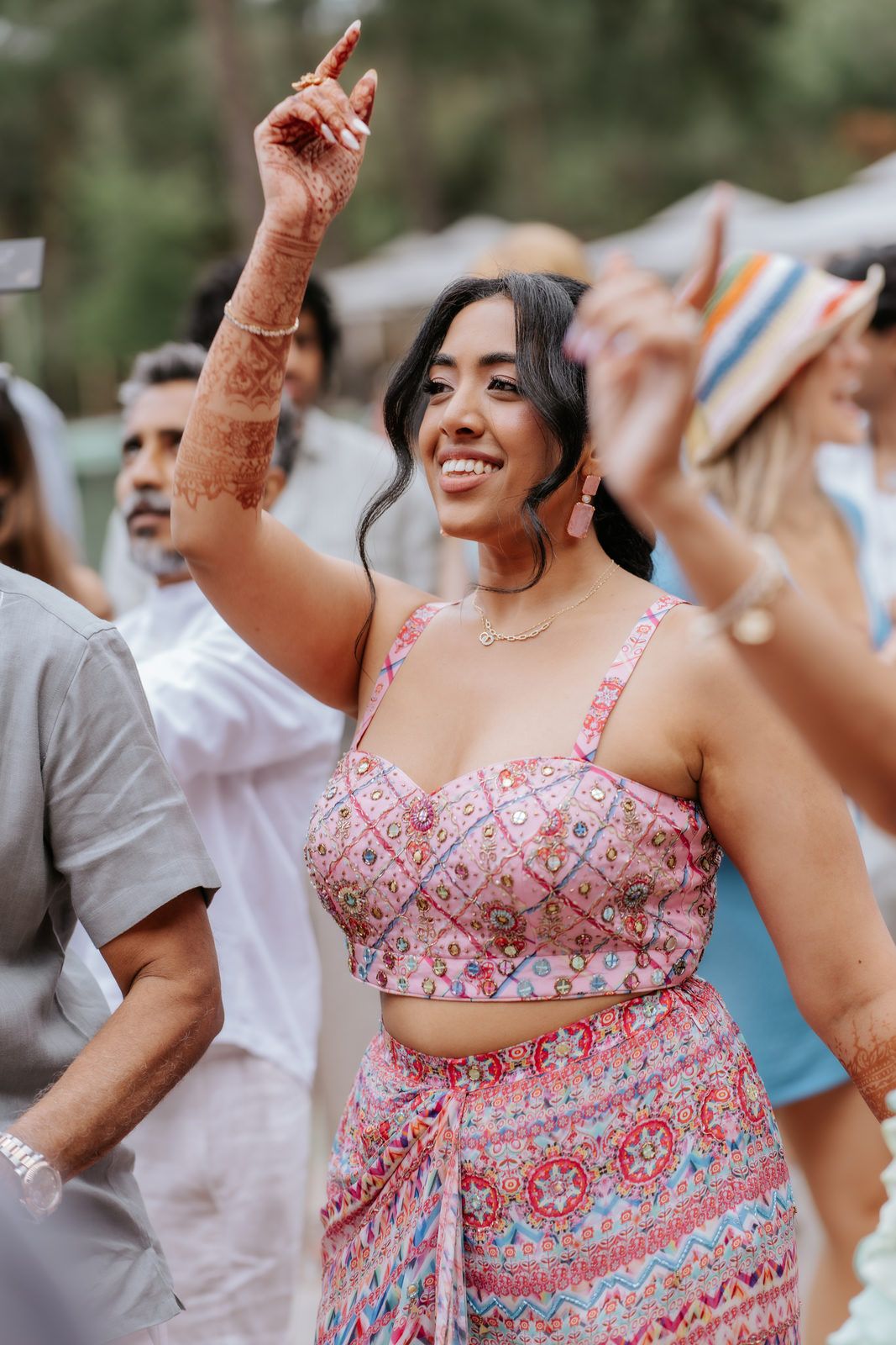 Woman in pink printed co-ord set dancing joyfully at an outdoor celebration with henna on her hand.