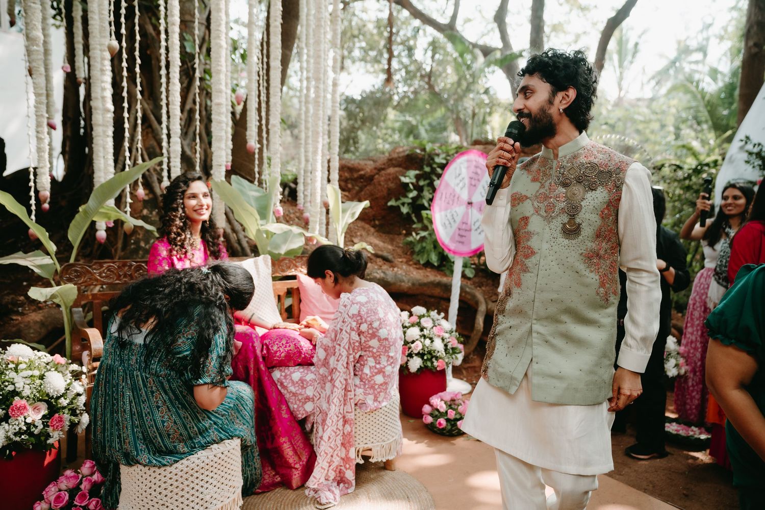 Groom in embroidered sherwani holds pink fan at outdoor Indian wedding ceremony surrounded by guests in colorful attire.