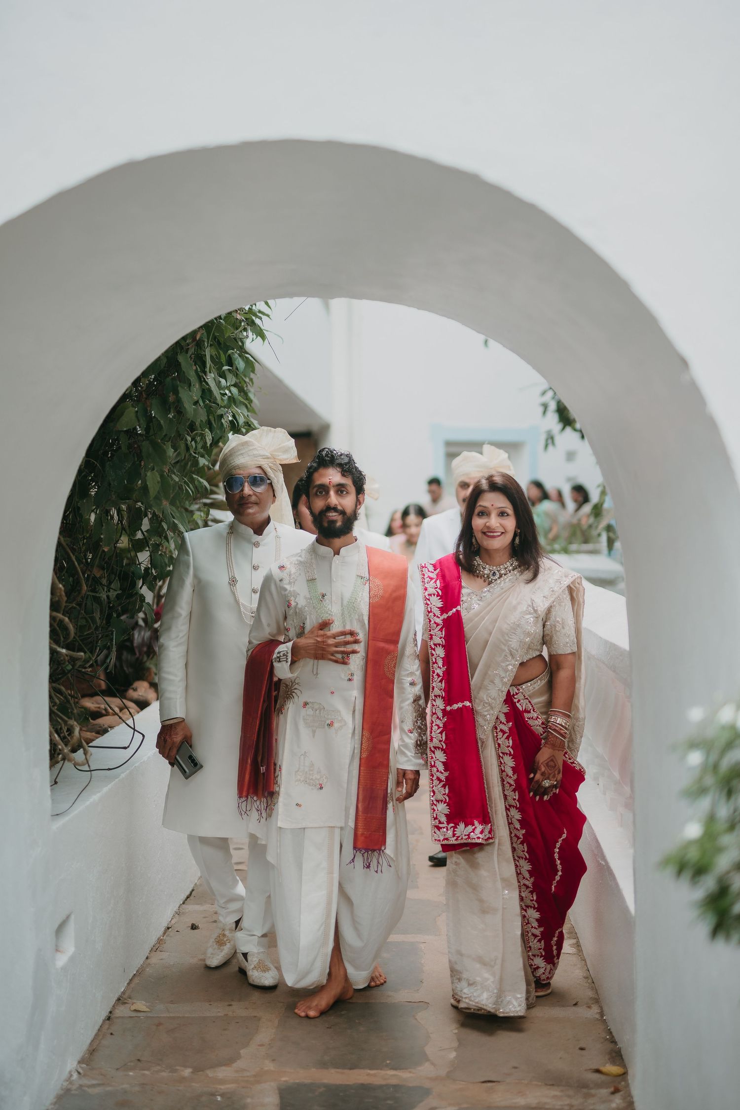 Indian wedding procession with groom in white sherwani and red dupatta walking through white arch with family.