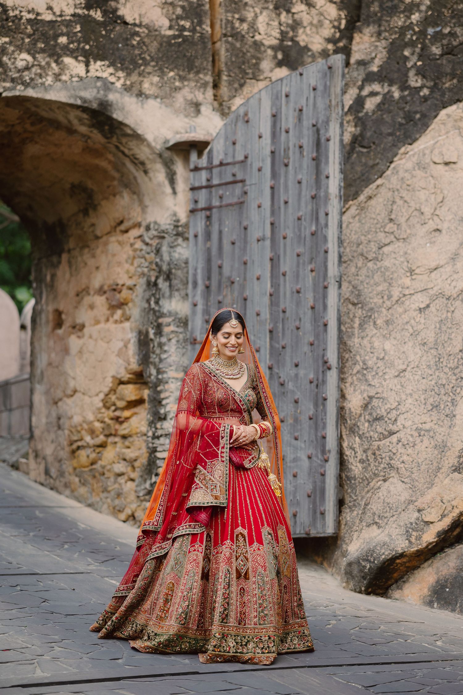 Bride in red embroidered lehenga posing against rustic stone wall with wooden door.