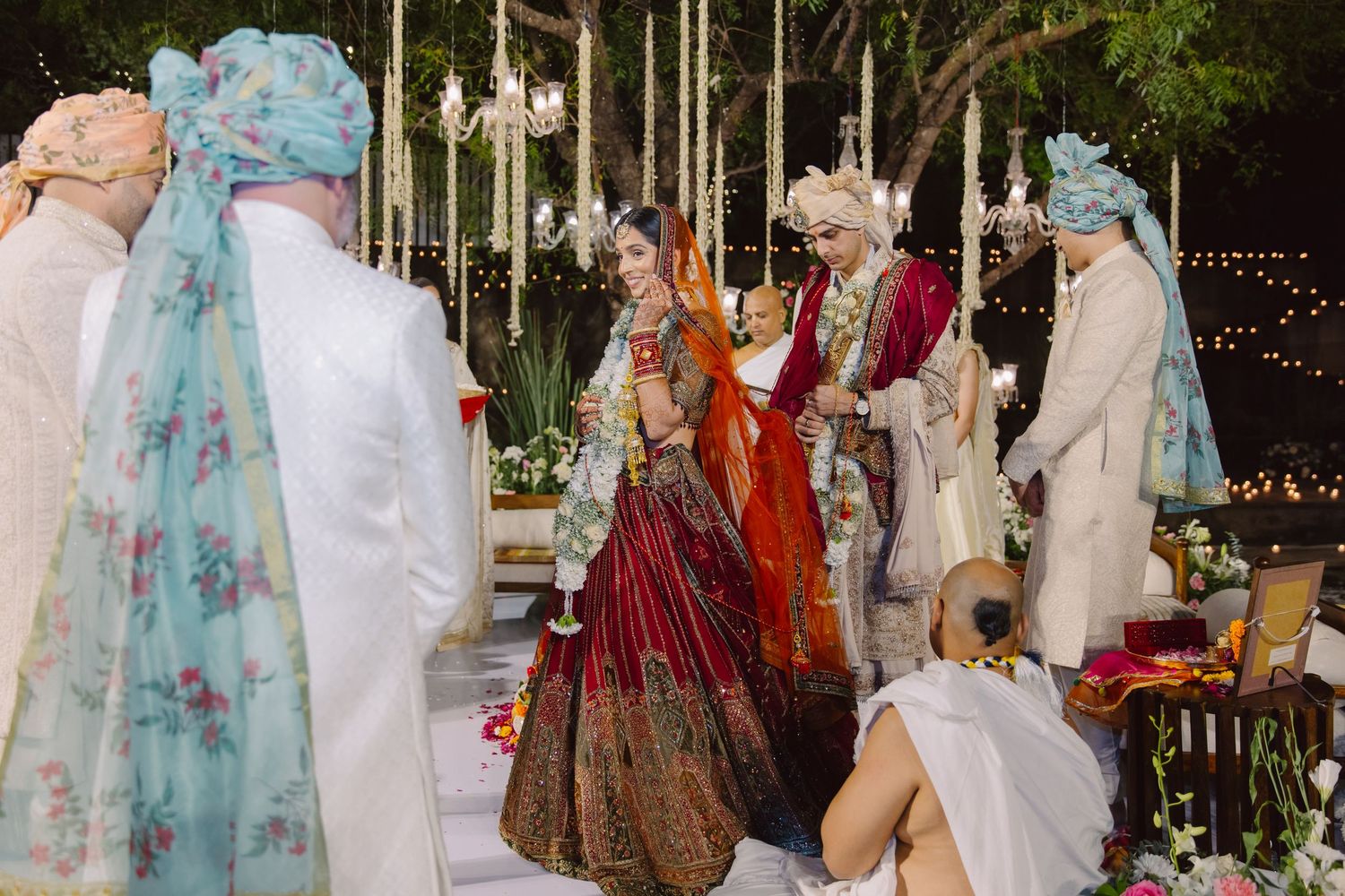 Traditional Indian wedding ceremony with guests gathered around floral decorations and string lights outdoors.