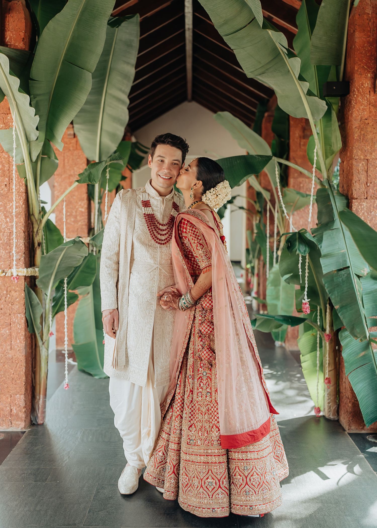 A couple in traditional Indian wedding attire poses in a tropical garden setting with banana plants.