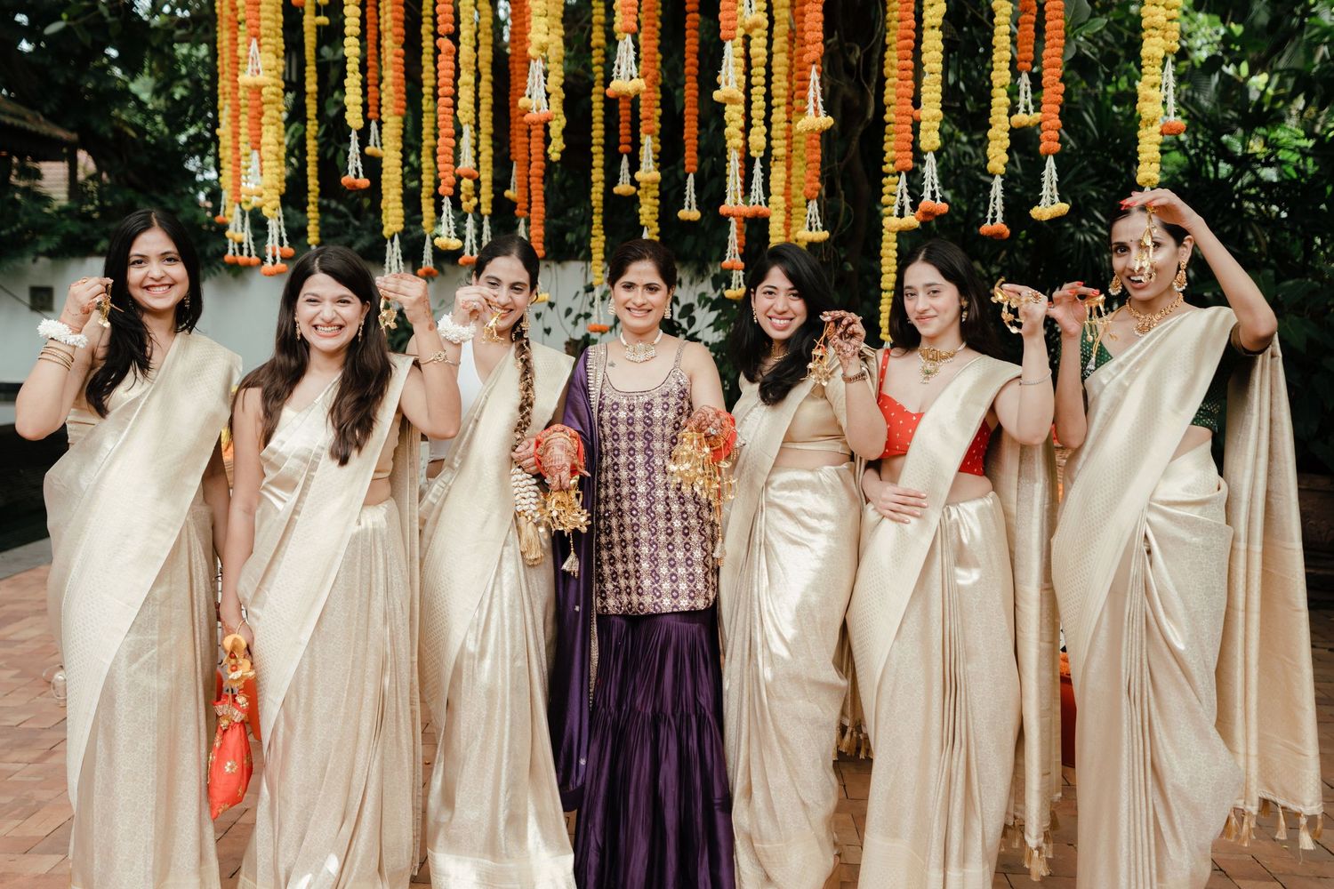 Seven women in cream sarees pose joyfully at an Indian wedding celebration with vibrant marigold floral decorations.
