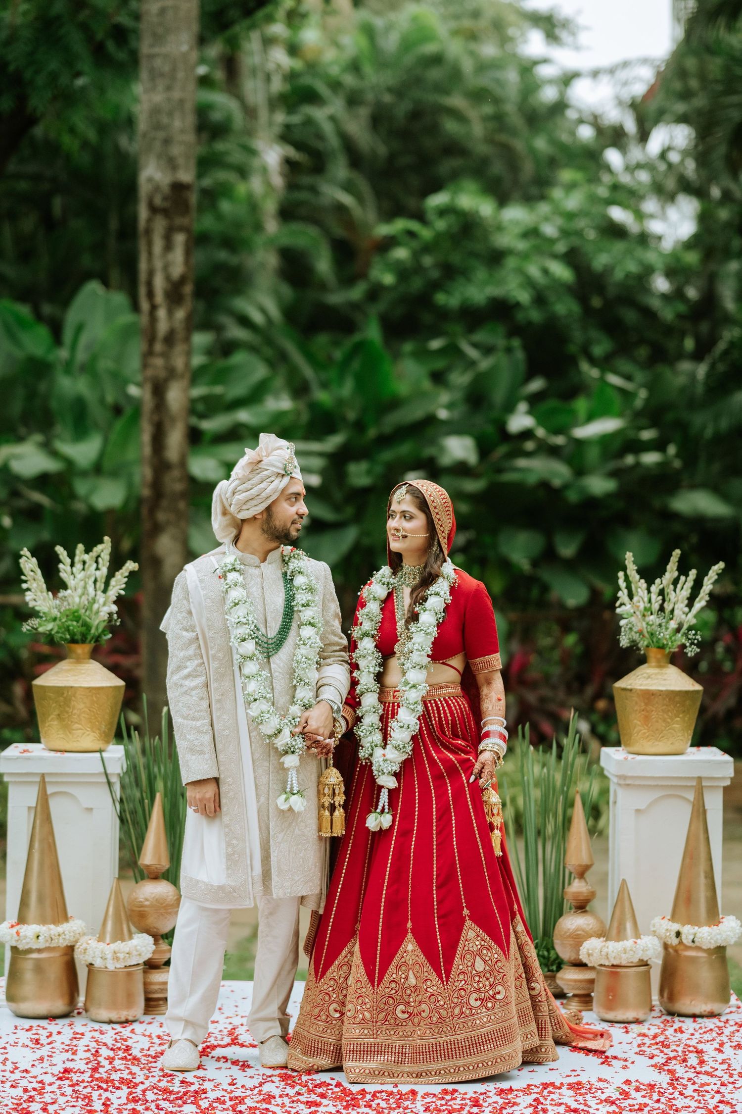 Indian bride in red lehenga and groom in white sherwani exchange garlands at outdoor Hindu wedding ceremony.