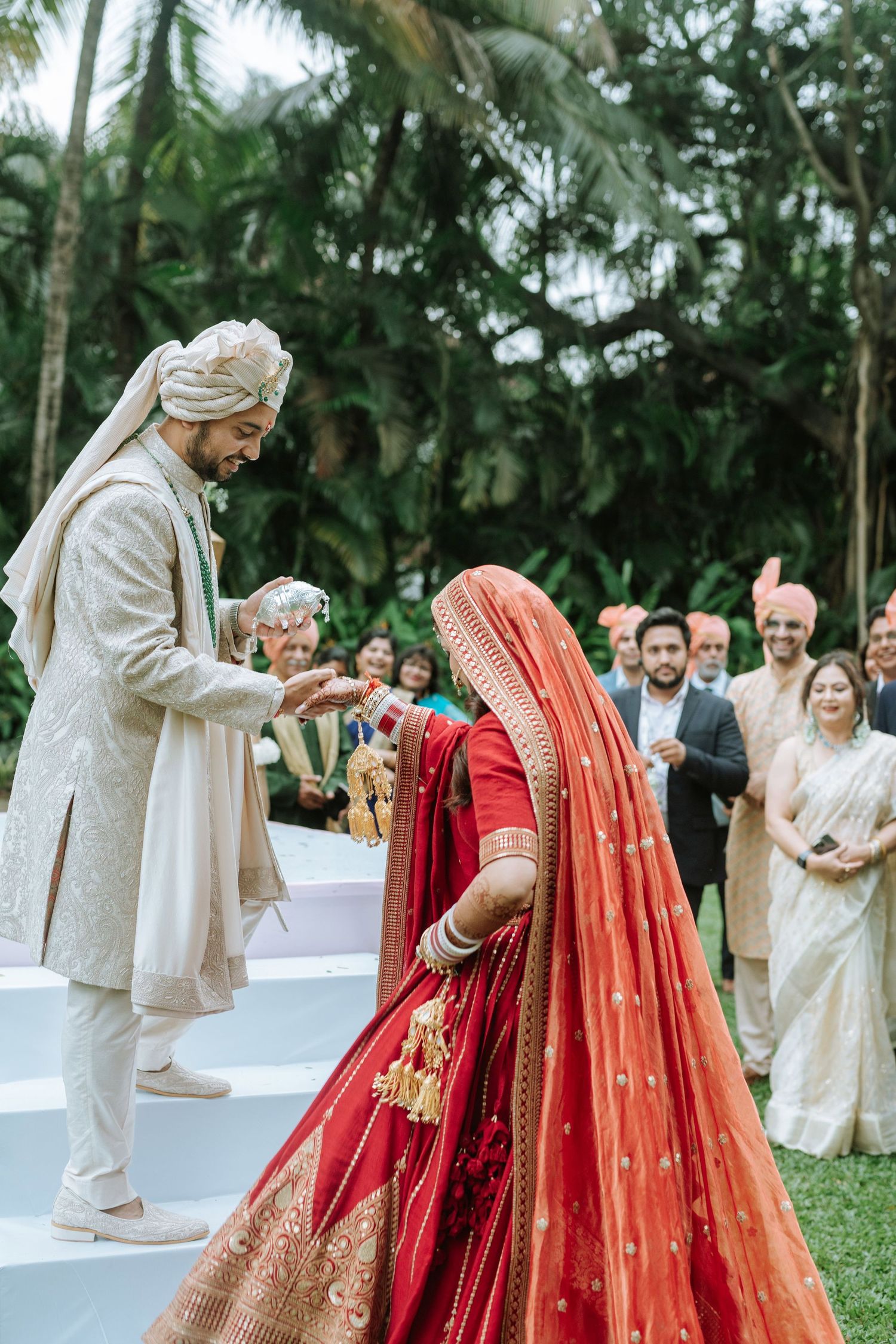 Groom in white sherwani welcomes bride in red lehenga at outdoor Indian wedding ceremony with guests watching.