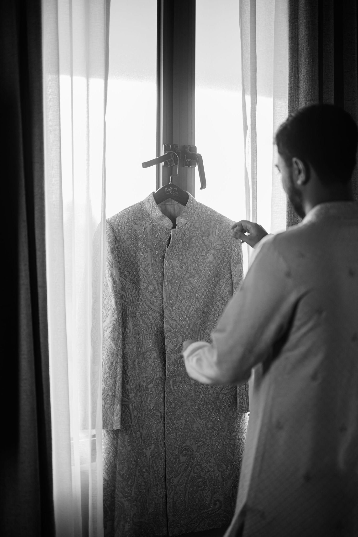 Black and white photo of a groom examining his sherwani hanging by a window before his wedding ceremony.
