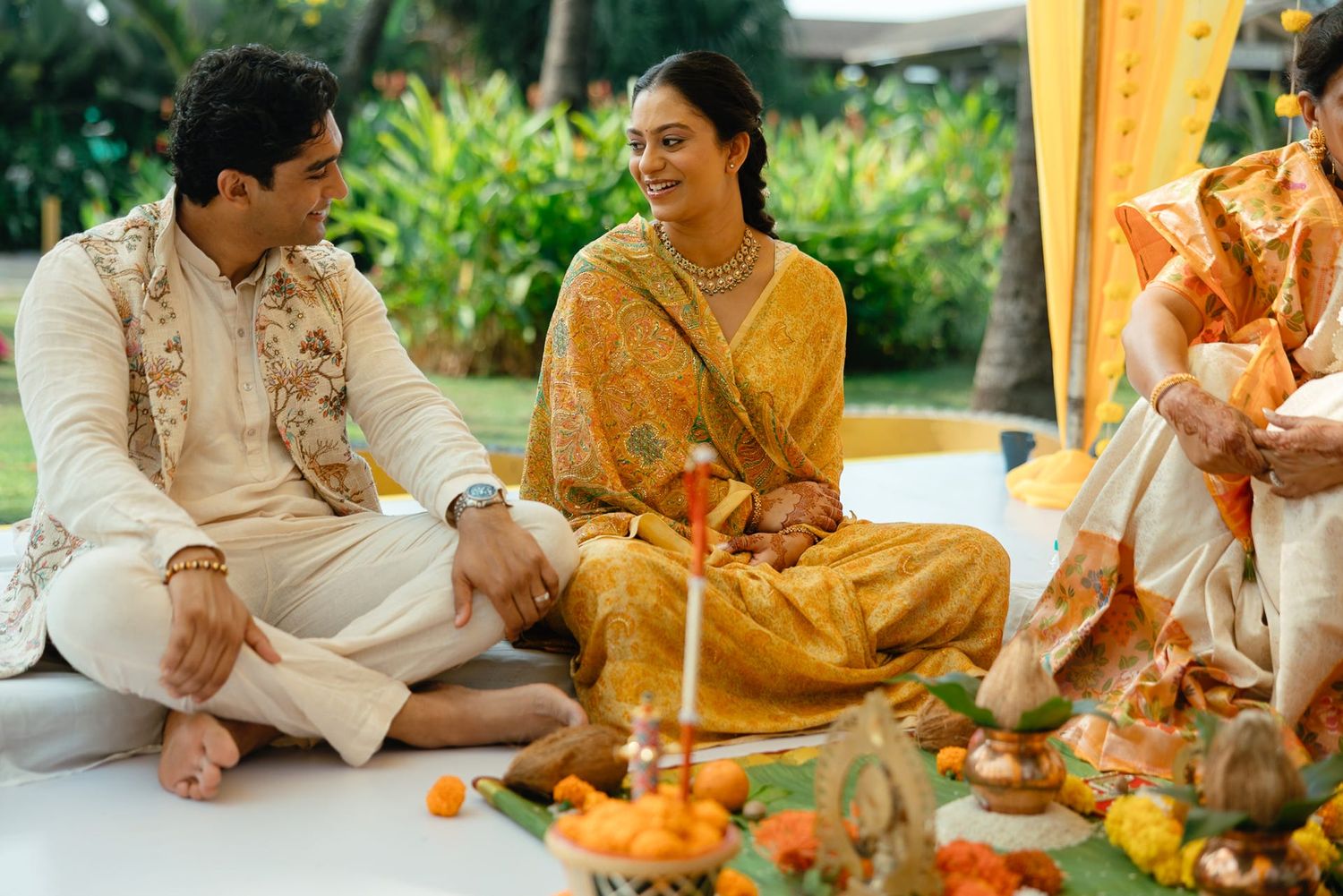 Couple in traditional Indian wedding attire sitting together smiling during a Hindu ceremony with floral decorations.