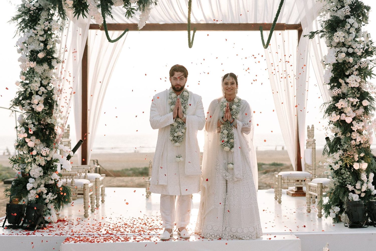 Couple in white traditional attire standing under floral arch with rose petals falling during outdoor wedding ceremony.