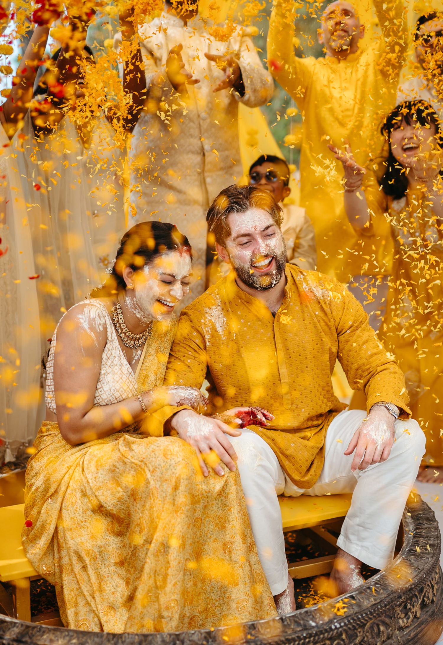 Couple laughing during a vibrant Indian haldi ceremony, covered in yellow turmeric powder with guests celebrating behind them.
