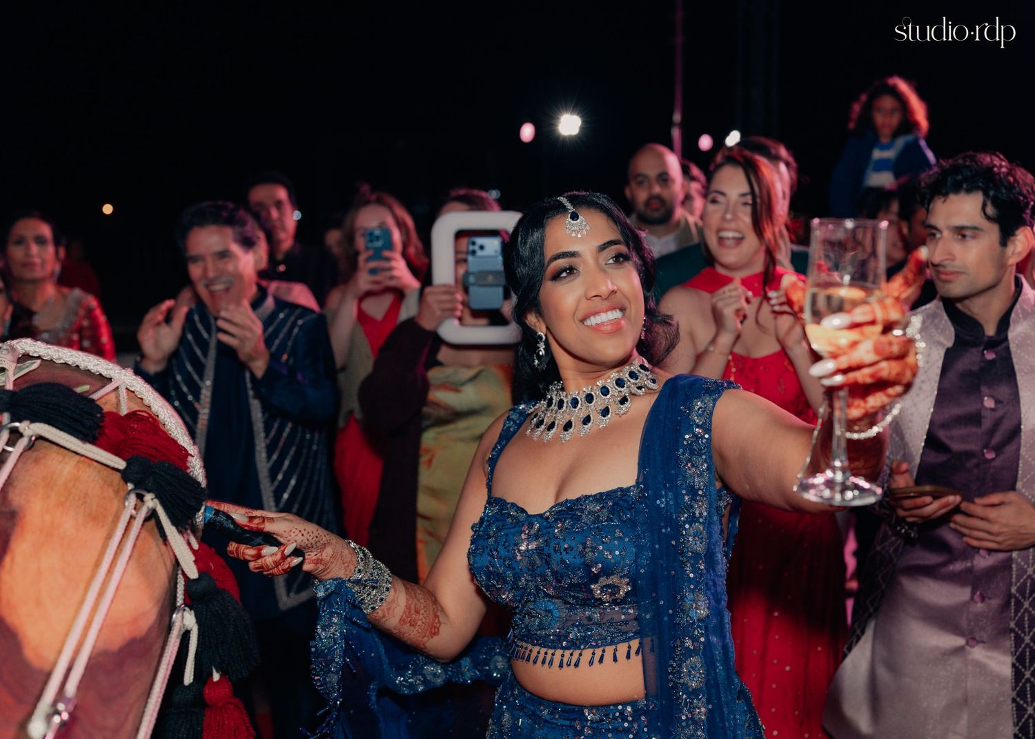 Woman in blue lehenga dancing joyfully at Indian wedding celebration with dhol drummer and guests.