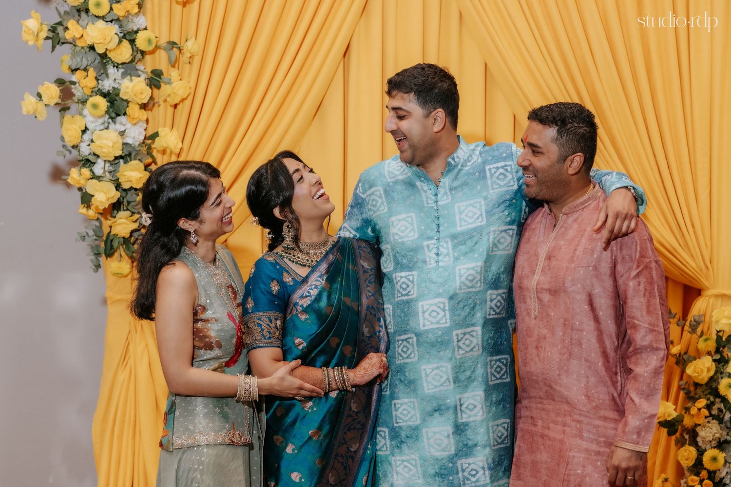 Family members in colorful Indian attire laugh together at a festive celebration with yellow drapes and floral decor.