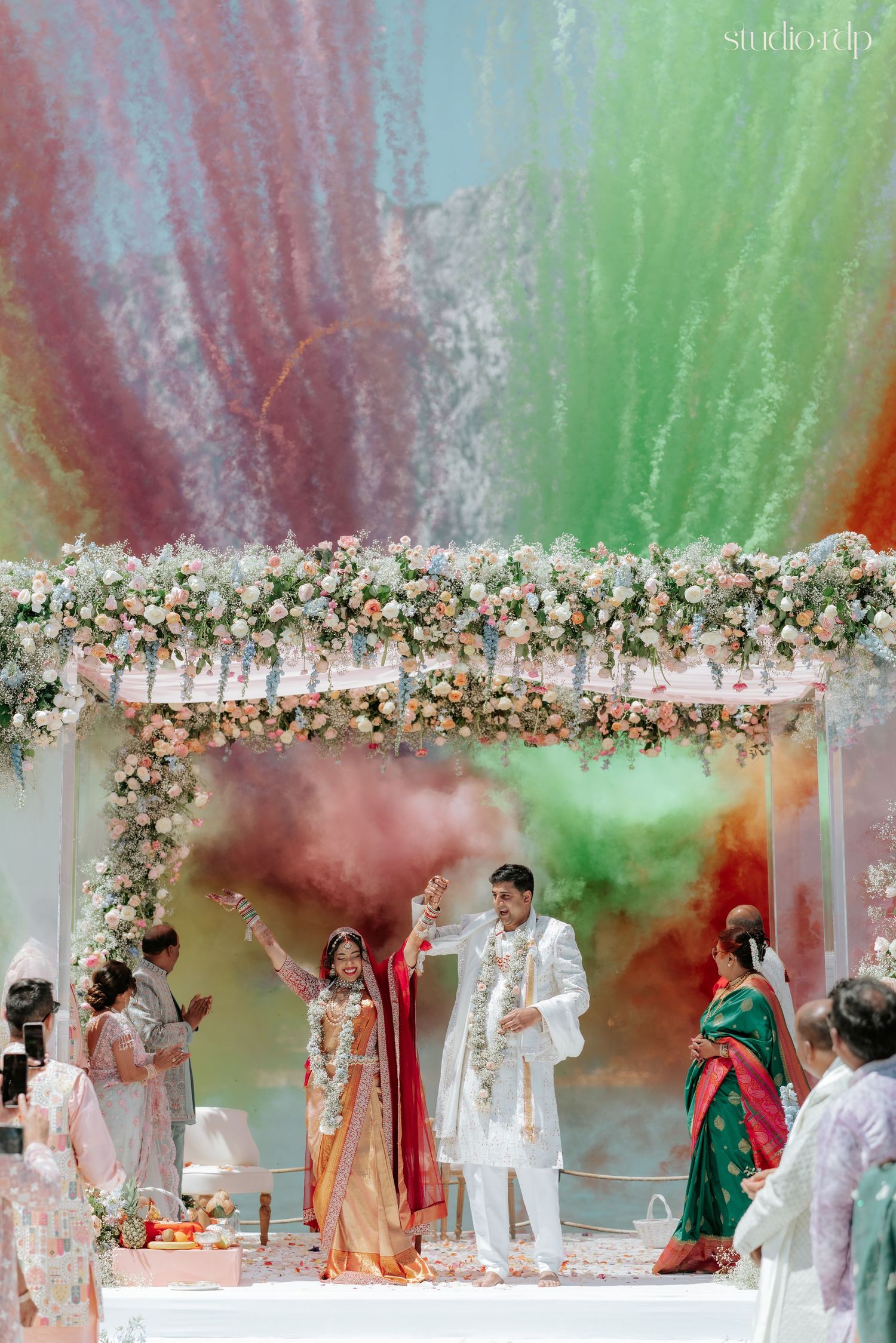 Indian wedding couple celebrates under floral mandap with vibrant Holi color powder burst in pink, green, and orange.