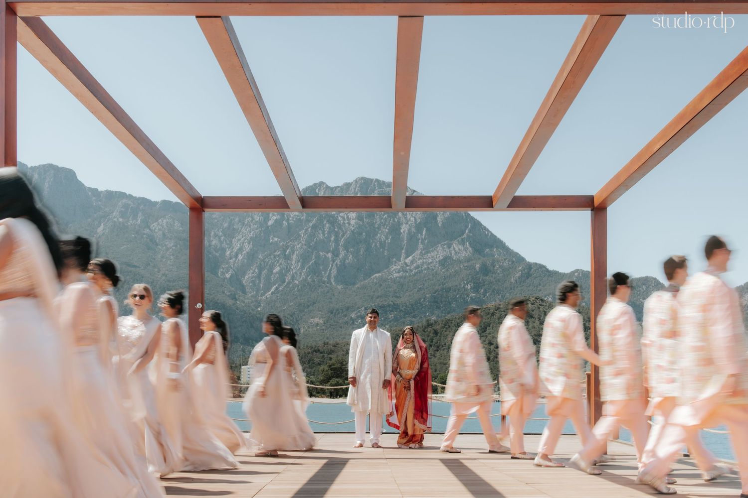 Indian wedding party in blush and white attire walks under pergola with mountain lake backdrop.