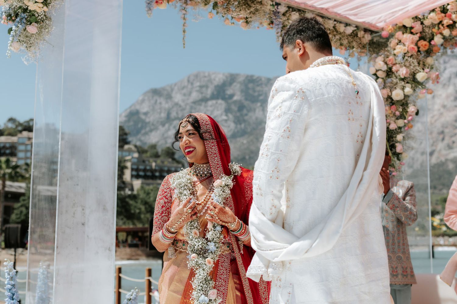 Indian bride and groom exchange vows at an outdoor wedding ceremony with mountain and floral backdrop.