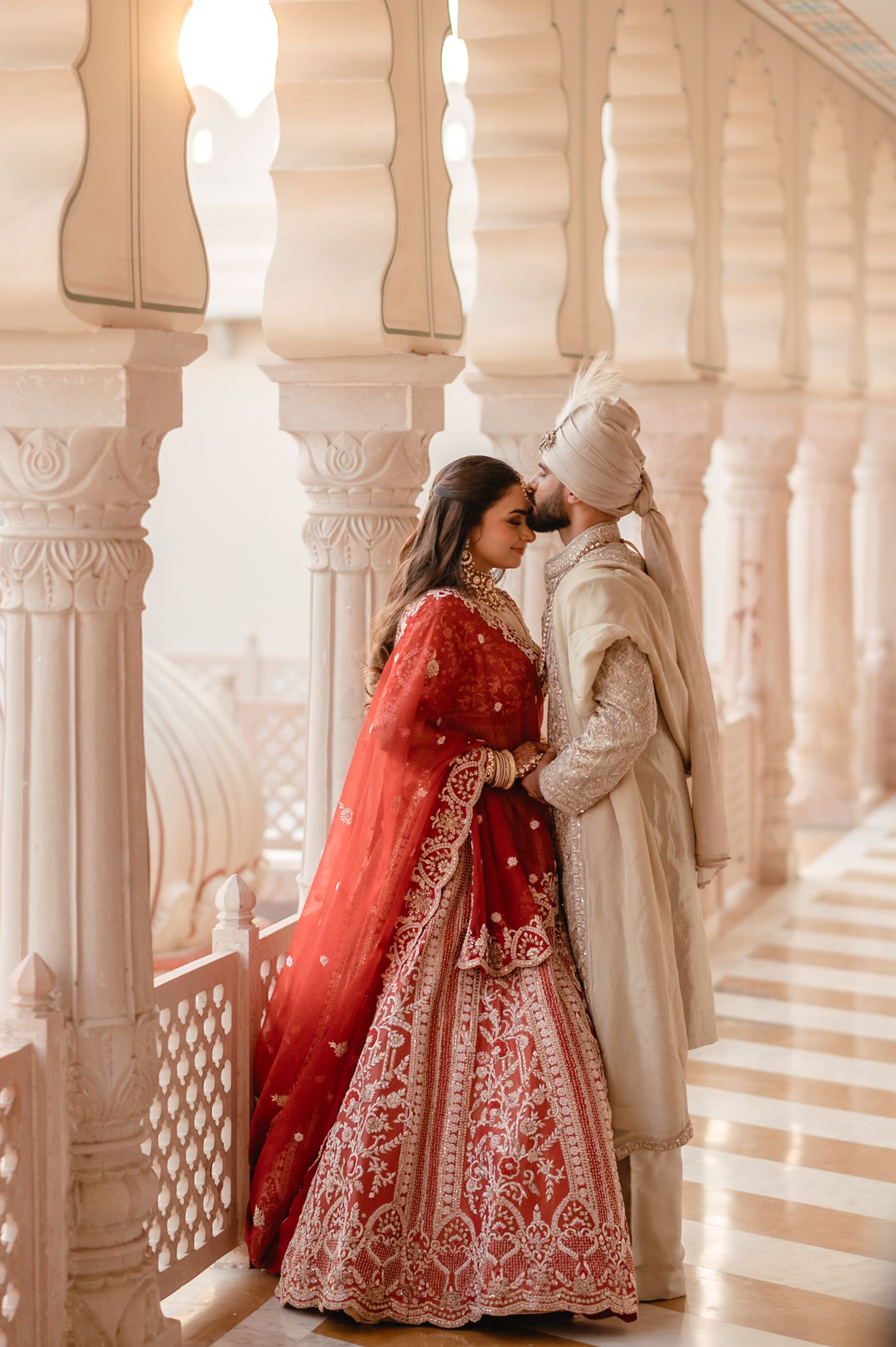 Indian bride in red lehenga and groom in white sherwani share romantic moment among ornate marble columns.