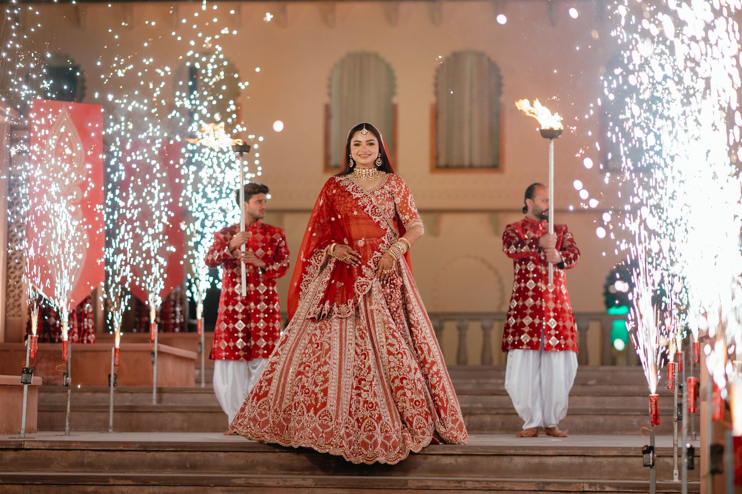 Bride in red embroidered lehenga walks down steps amid sparkling fireworks at a grand Indian wedding ceremony.
