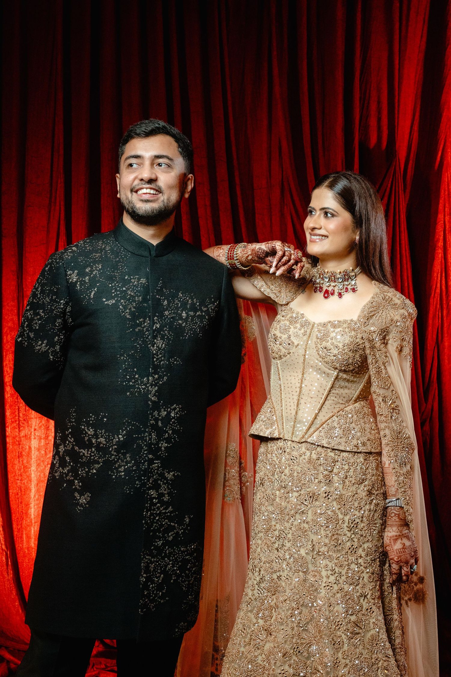Indian couple in traditional wedding attire, man in black sherwani and woman in gold lehenga, posing before red curtain.