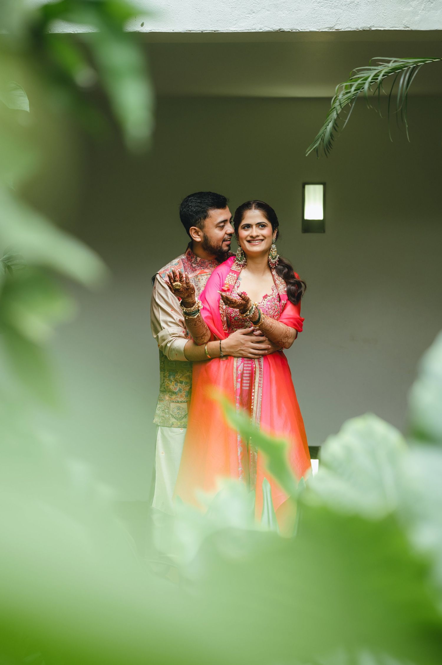 Couple in traditional Indian attire embracing outdoors, woman in pink lehenga, man in gold kurta, lush green foliage framing.