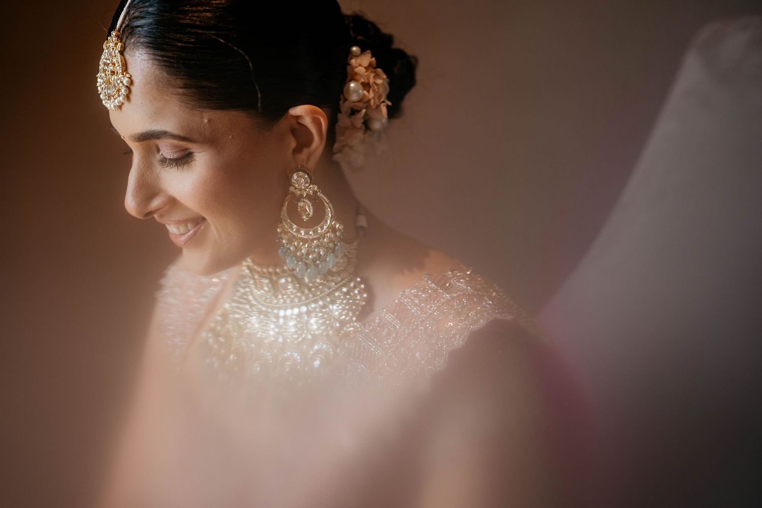 A bride in an elegant white gown and traditional jewelry poses in soft, romantic lighting at her wedding.