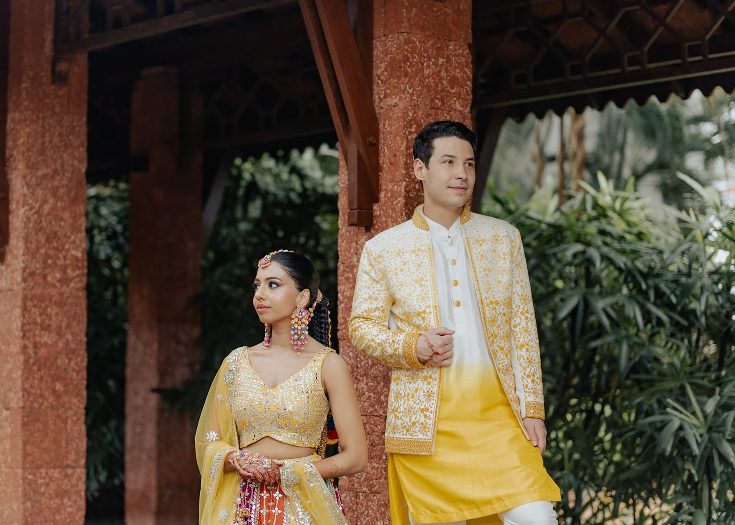 Couple in matching yellow traditional South Asian wedding attire posing outdoors near ornate wooden pillars.