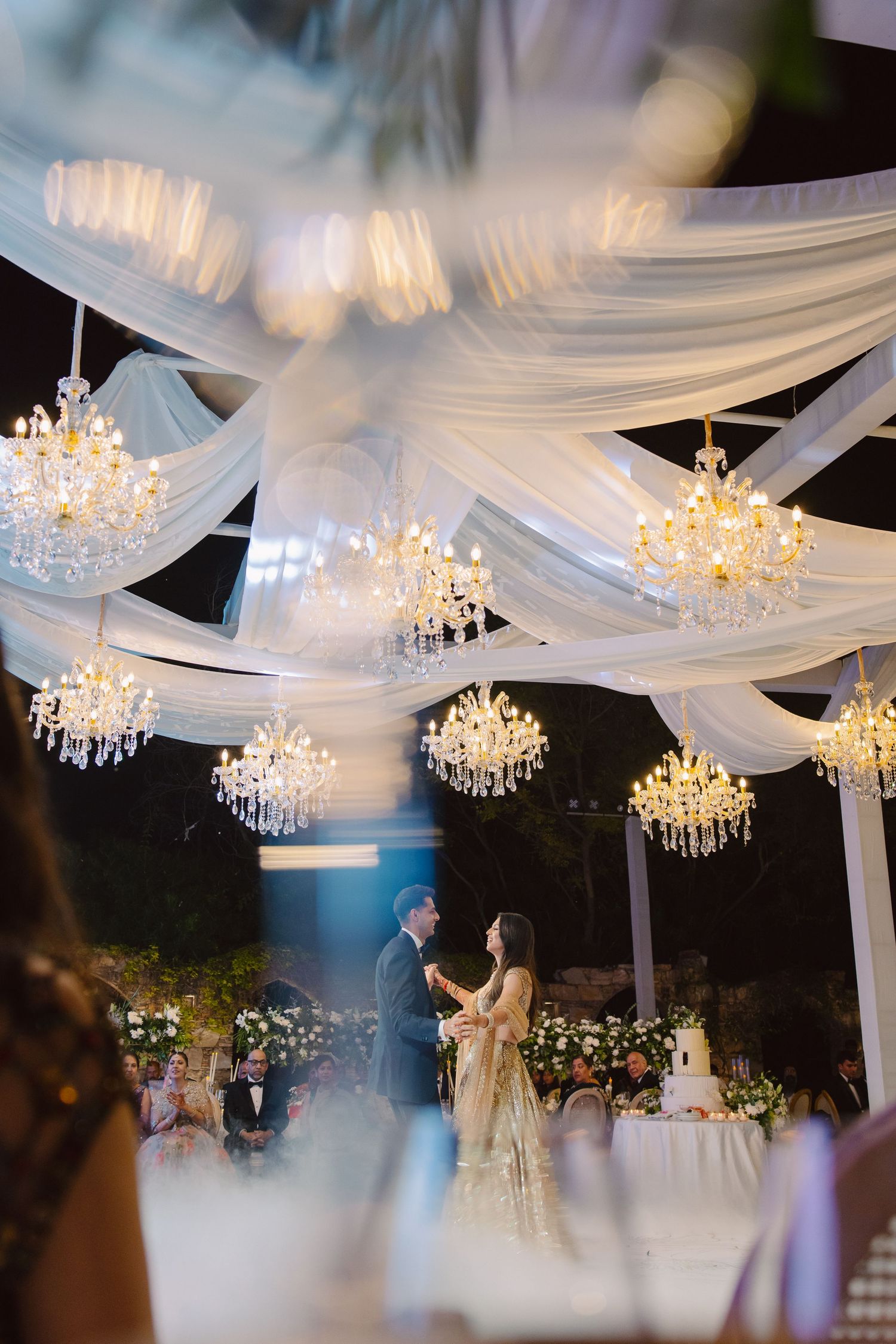 Elegant outdoor wedding reception with white fabric draping and crystal chandeliers overhead.