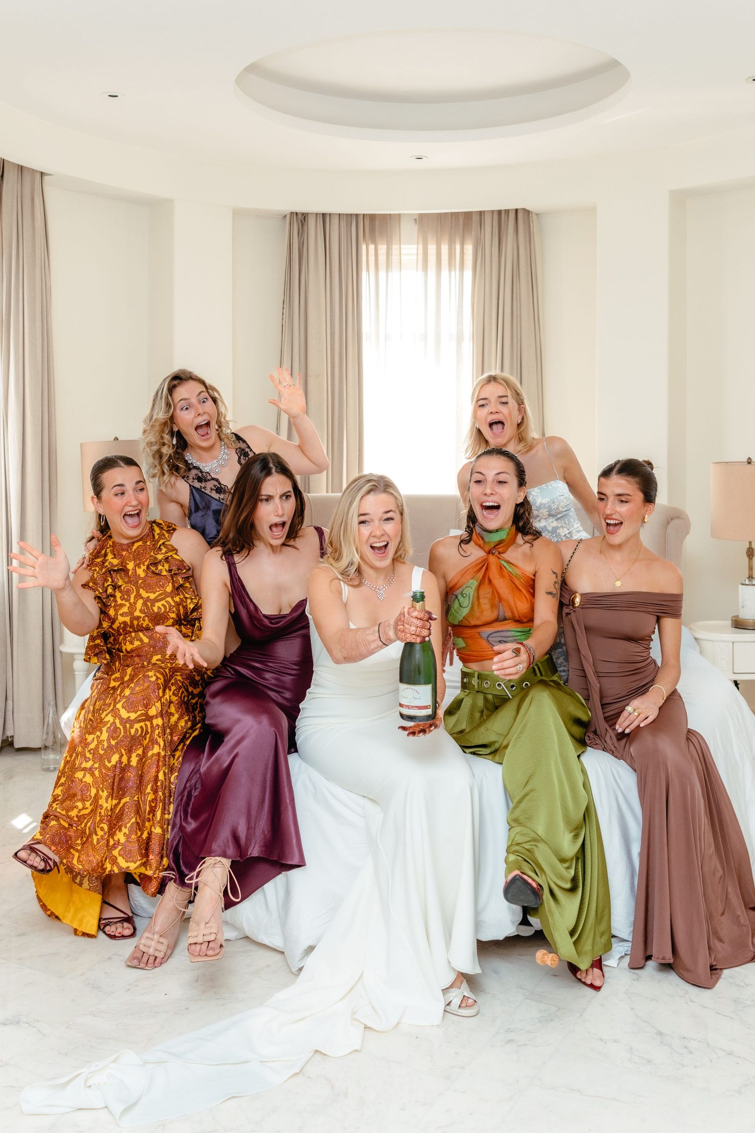 Bridesmaids in colorful dresses celebrate joyfully with champagne in a bright modern room.