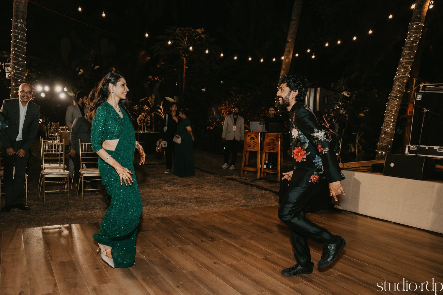 A couple performs an energetic dance routine on a wooden floor under twinkling string lights at an evening reception.