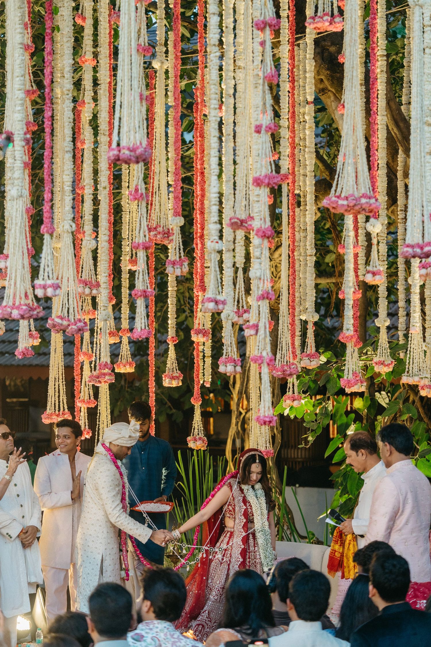 Indian wedding ceremony under floral garland backdrop with bride in red lehenga and groom in white sherwani.