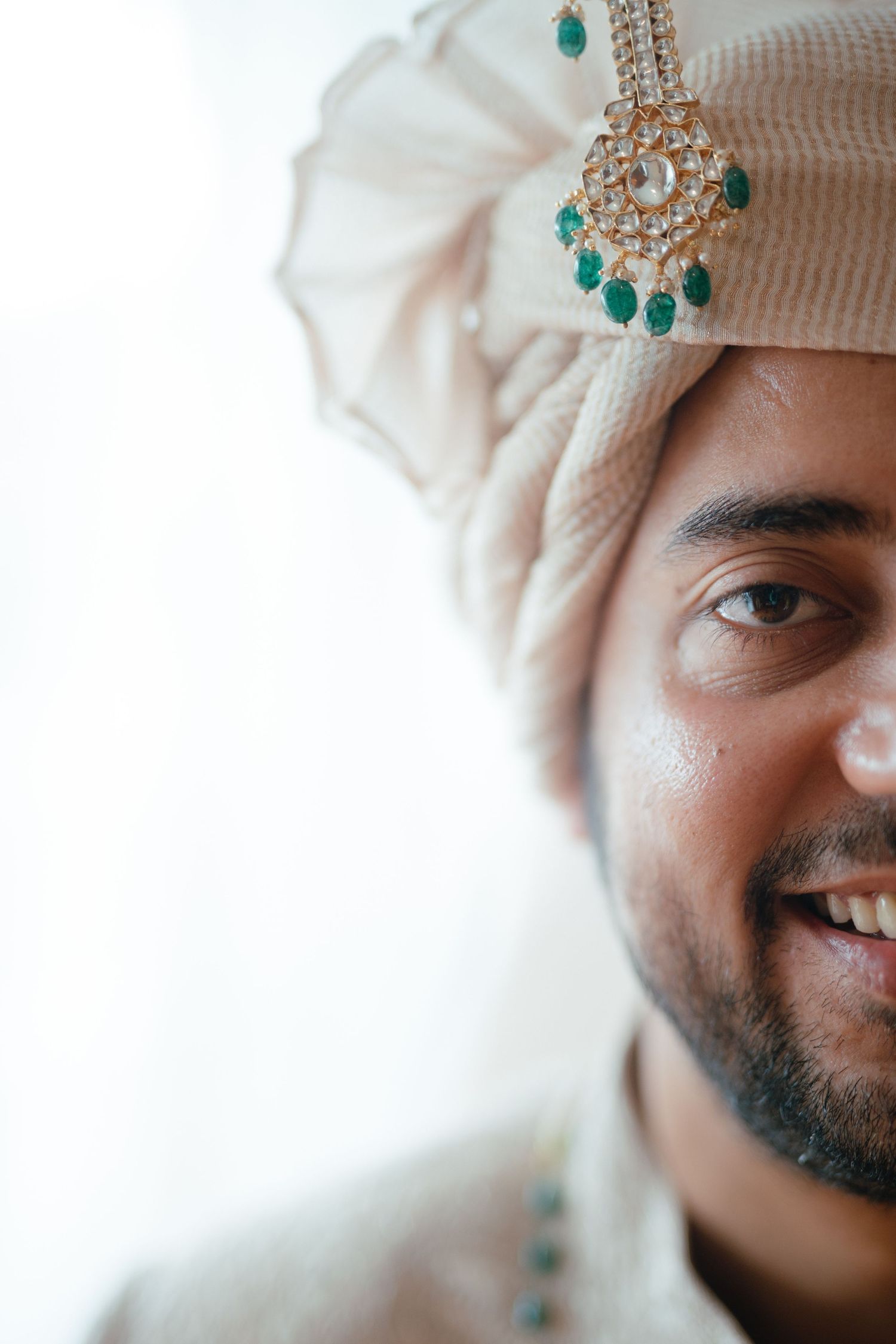 Smiling Indian groom wearing traditional cream turban with gold and emerald jeweled brooch at wedding ceremony.