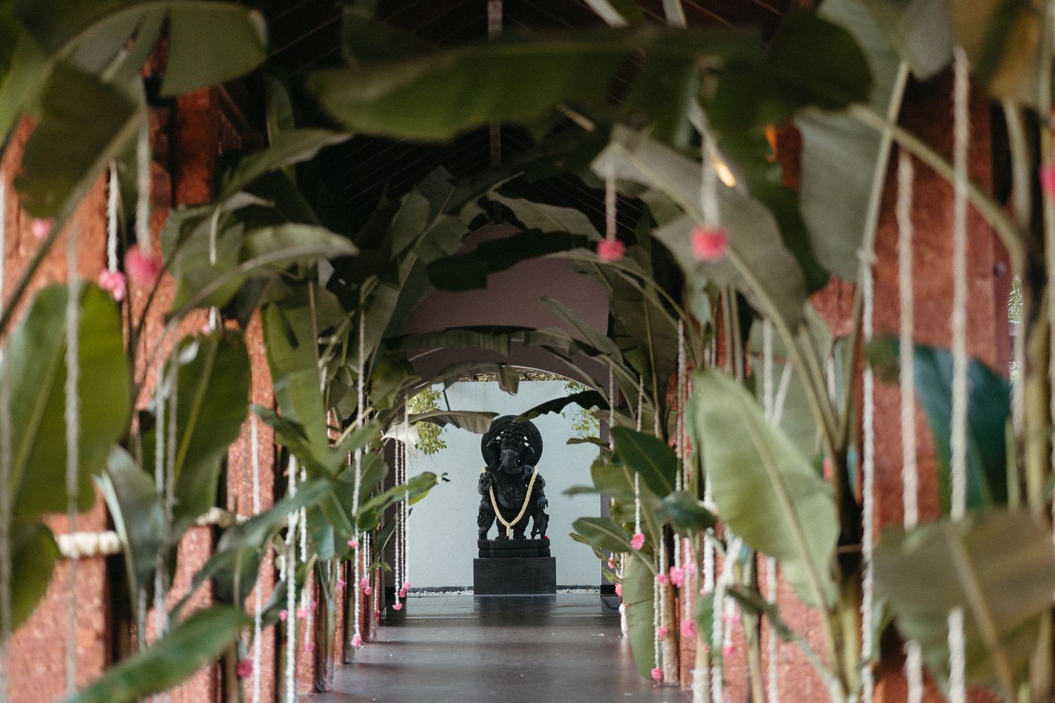 A lush tropical corridor with arching banana plants framing a dark statue at the end of a brick hallway.