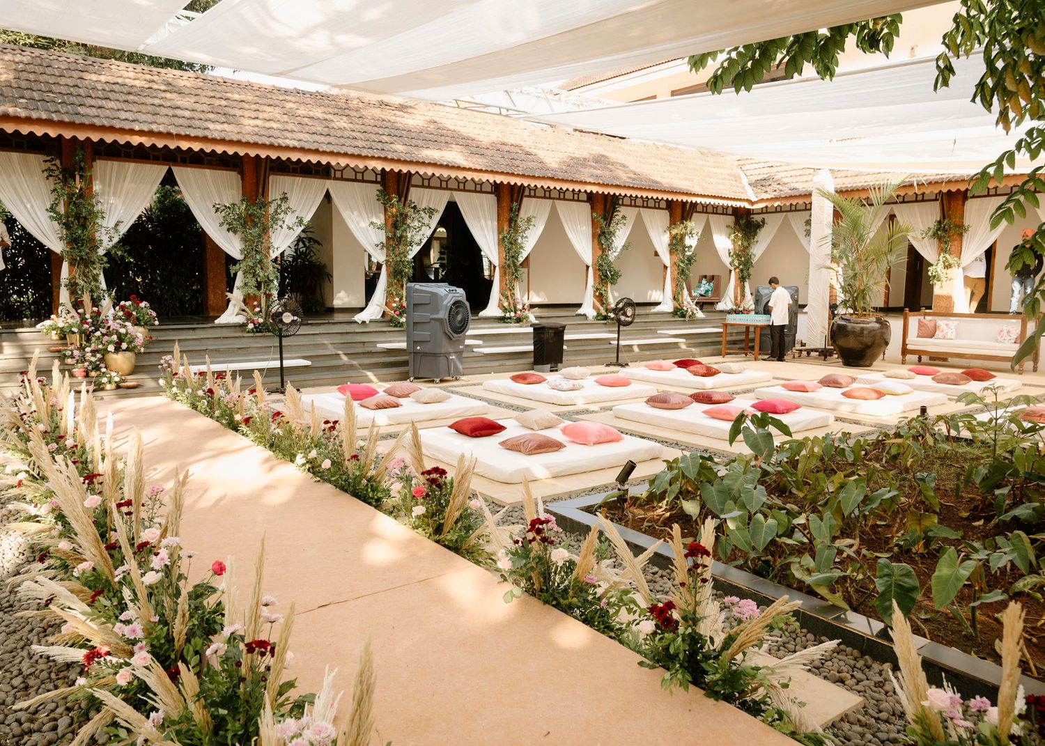 Elegant outdoor wedding venue with white cushioned seating, floral arrangements, and draped canopy in a courtyard setting.