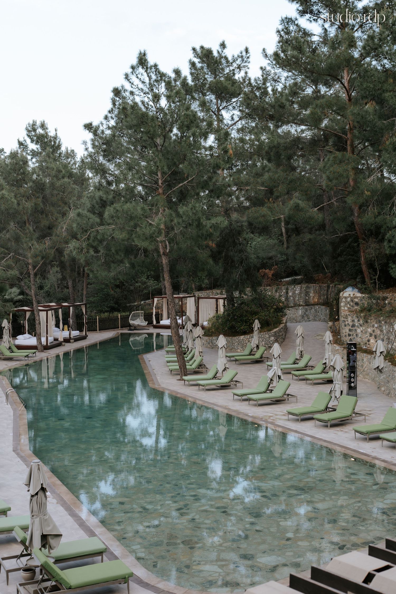 Luxury resort pool with green lounge chairs, closed umbrellas, and pine trees reflected in calm turquoise water.