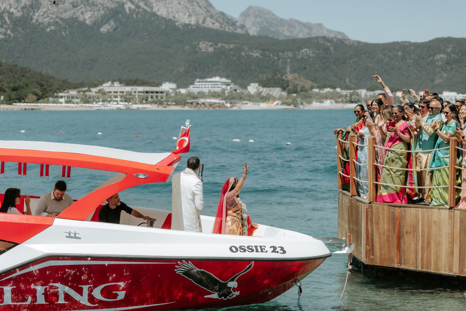 Indian wedding guests wave from a wooden dock as a red speedboat named Ossie 23 departs on turquoise waters with mountains behind.