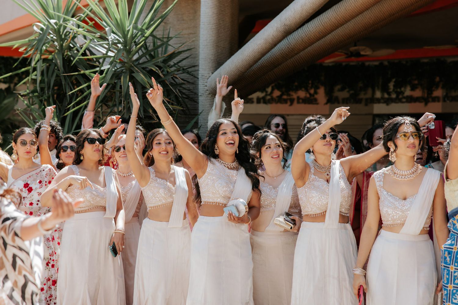 Bridesmaids in white dresses raise their arms joyfully during an outdoor Indian wedding celebration.