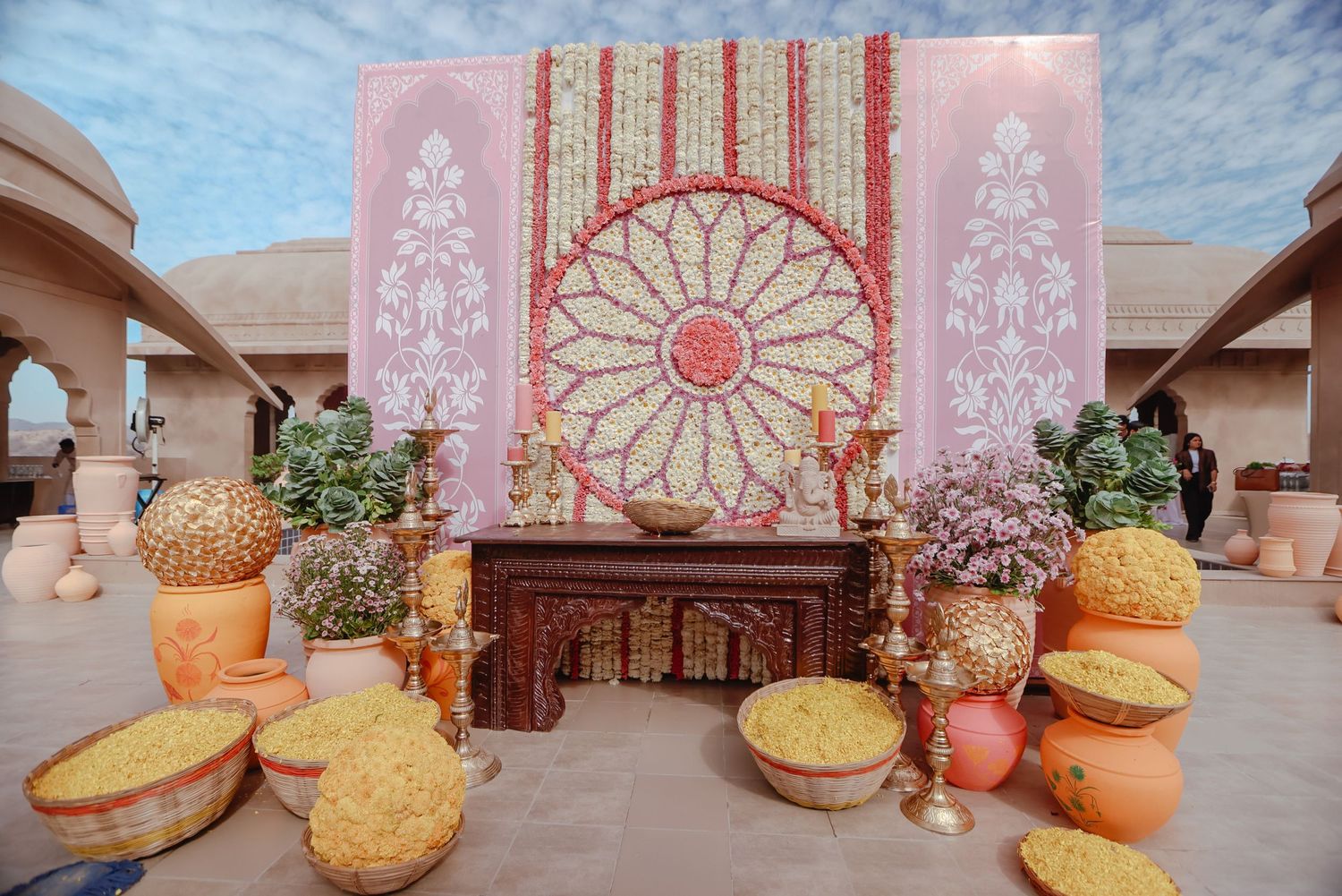 Pink and gold wedding ceremony backdrop with ornate mandala design and floral arrangements in rustic clay pots.
