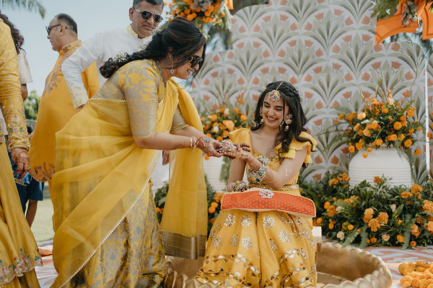 A group celebrates at a festive Indian wedding ceremony decorated with vibrant yellow and orange floral arrangements.