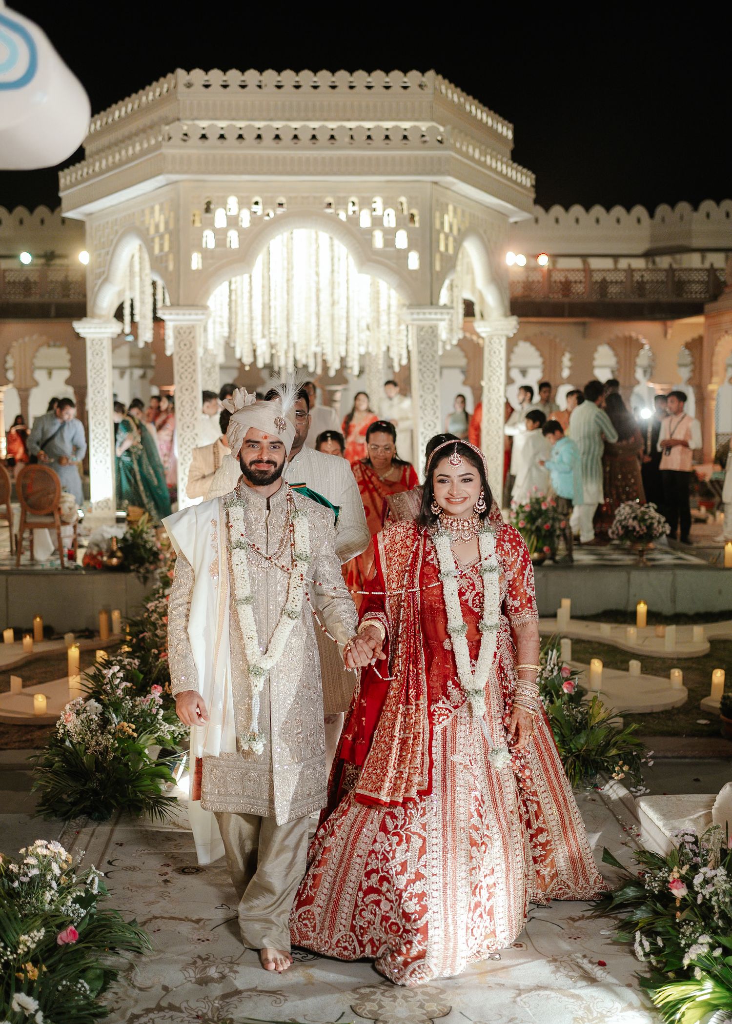Indian bride in red lehenga and groom in white sherwani walk together at an elegant outdoor wedding ceremony at night.